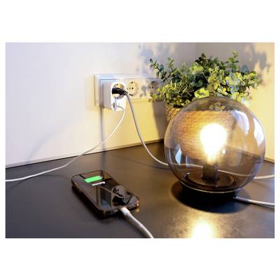 Smartphone charging on a desk beside a lit table lamp plugged into a wall outlet, with a potted plant in the background.