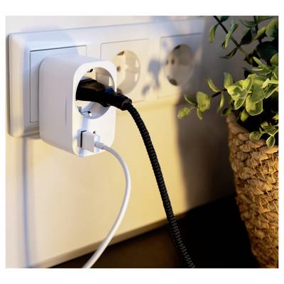 Close-up of a wall outlet with a white plug adapter and two charging cables, beside a potted plant.