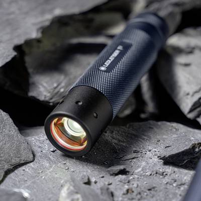 A flashlight with a glowing lens rests on dark, textured slate rocks. The flashlight has a black, textured grip handle.