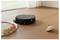 A black circular robot vacuum cleaning a wooden floor near a toy train on tracks and a spinning top in a sunlit room.