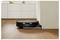 A black robotic vacuum cleaner is docking itself against a white baseboard in a room with wooden floors.