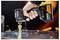 A hand holds a DeWalt cordless drill driving a gold stepped bit into a metal sheet, with sparks and metal shavings flying.