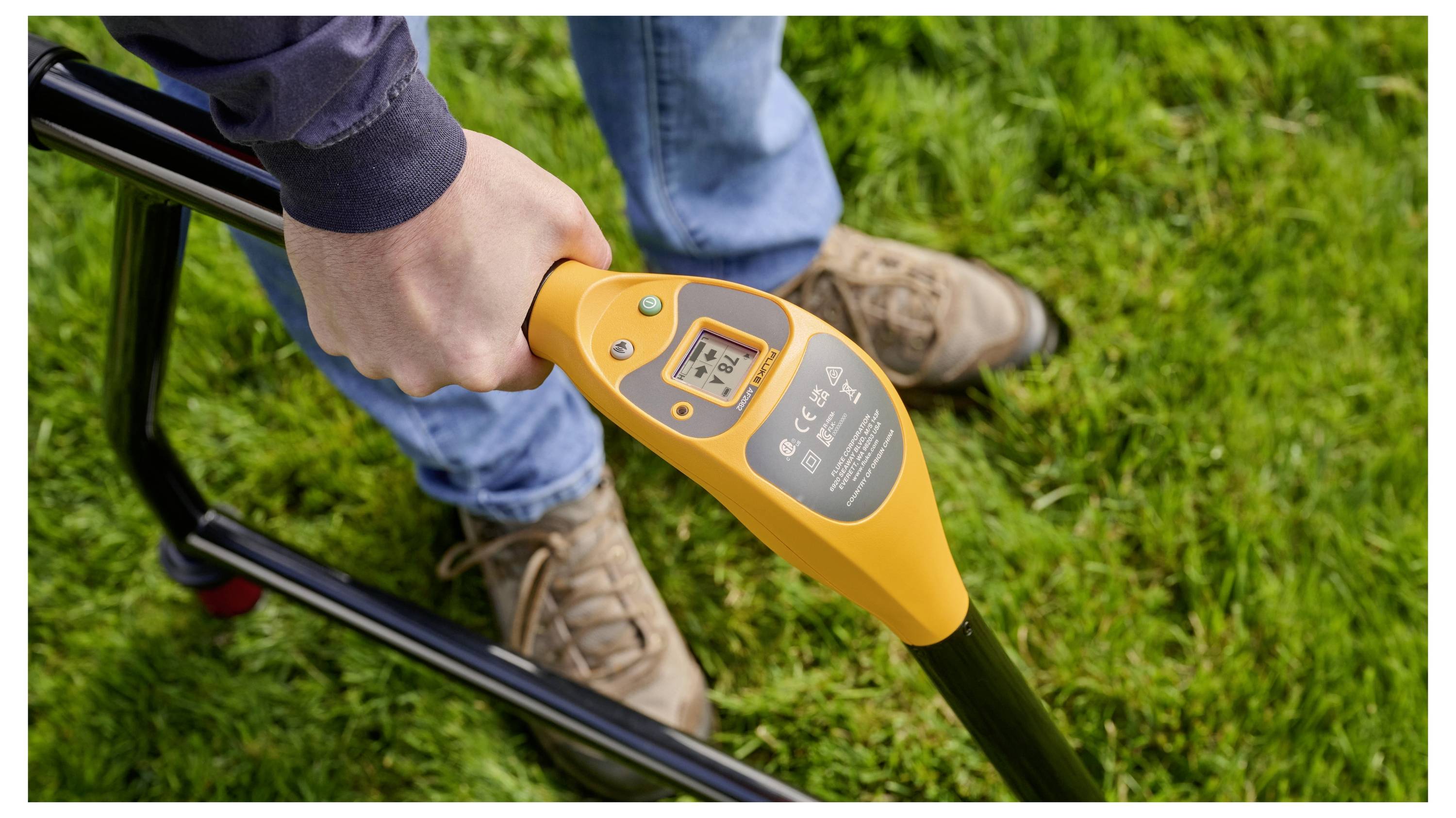 Close-up of a person using an orange electronic tool with a digital display on a black handle outdoors on grass.