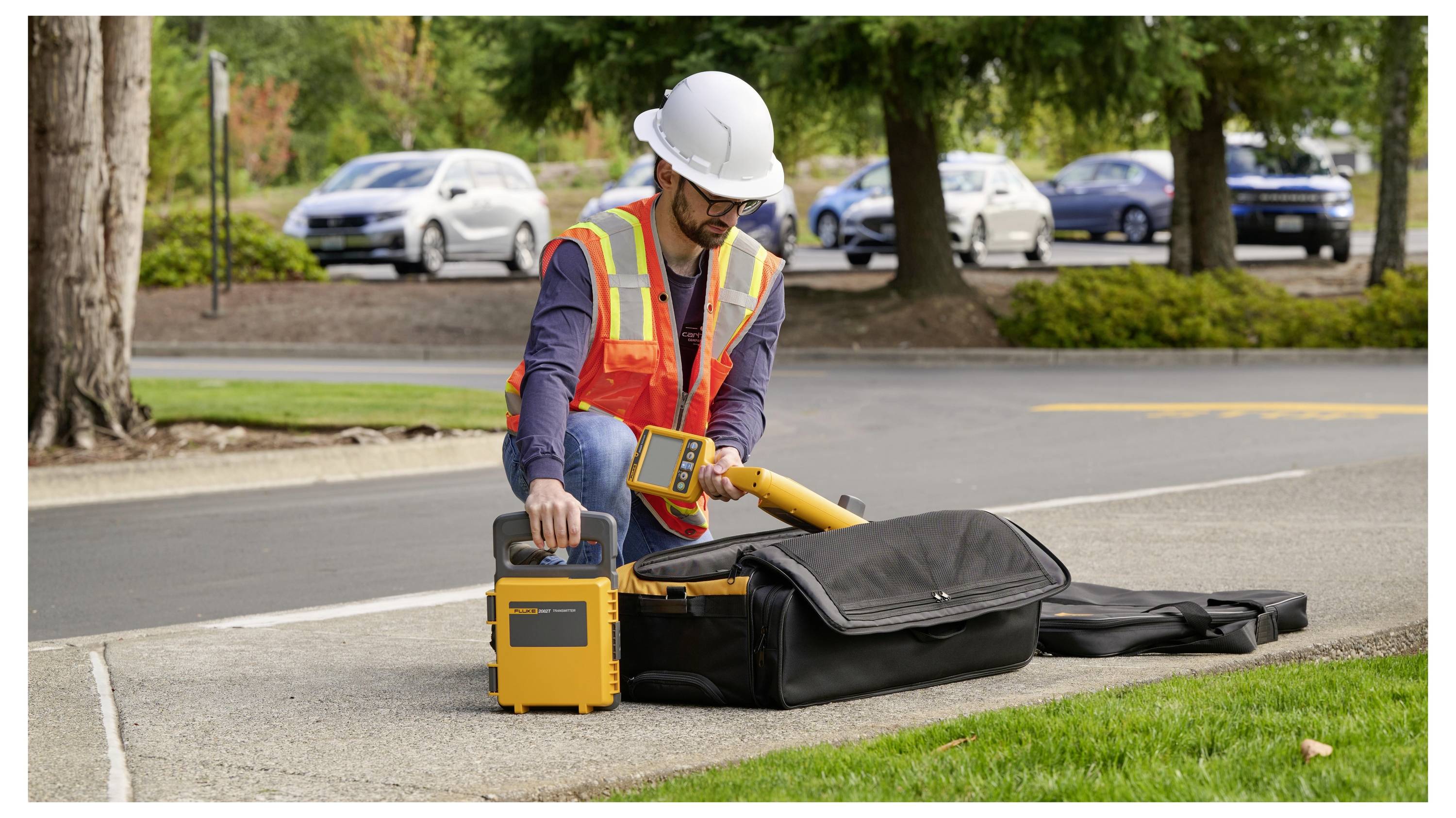 Utility worker in hard hat and safety vest operating roadside testing equipment beside an open case near the curb.
