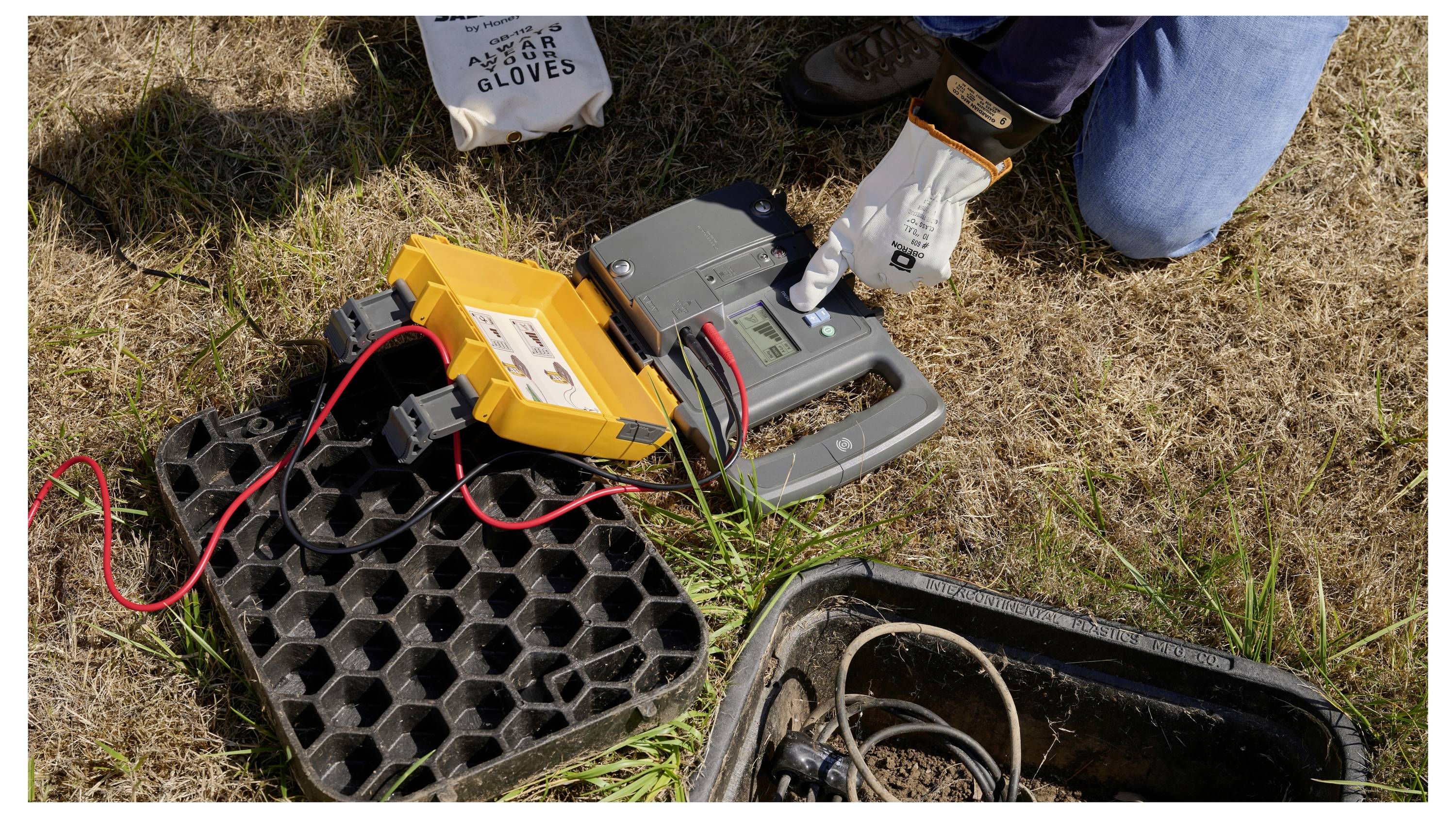 Person using gloves to connect a yellow battery tester/charger to a car battery outdoors near an open ground utility box<br>