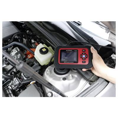 Hand holding an Autophix automotive scanner over a car engine bay, checking diagnostics near the battery and brake fluid reservoir.