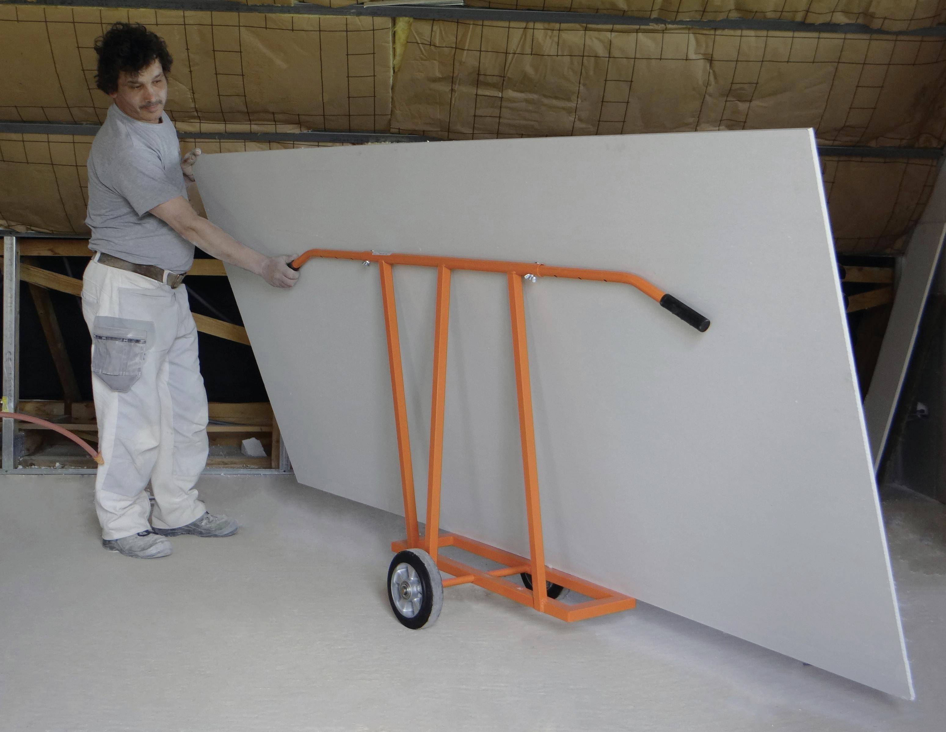 Worker using an orange drywall dolly to move a large white panel in a warehouse or workshop.