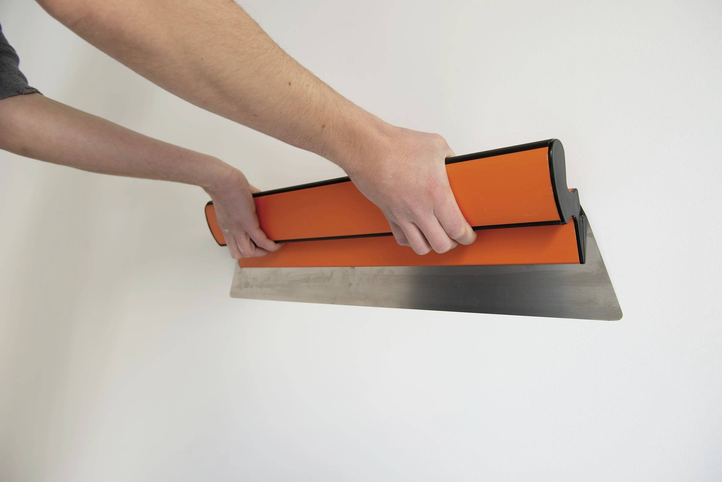 Hands holding a trowel against a white wall, applying or smoothing plaster with an orange-handled tool.