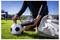Soccer player inflates a ball on the grass beside a sports bag, with another player practicing in the background.