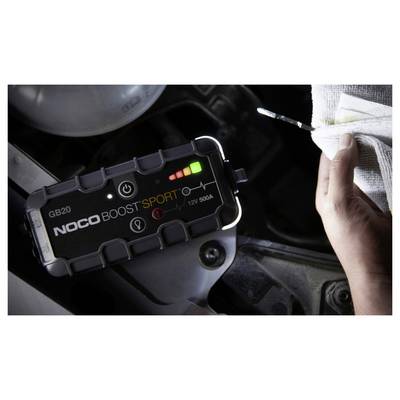 Portable NOCO Boost Sport jump starter connected in a car engine bay, with a hand holding jumper leads or cable nearby.