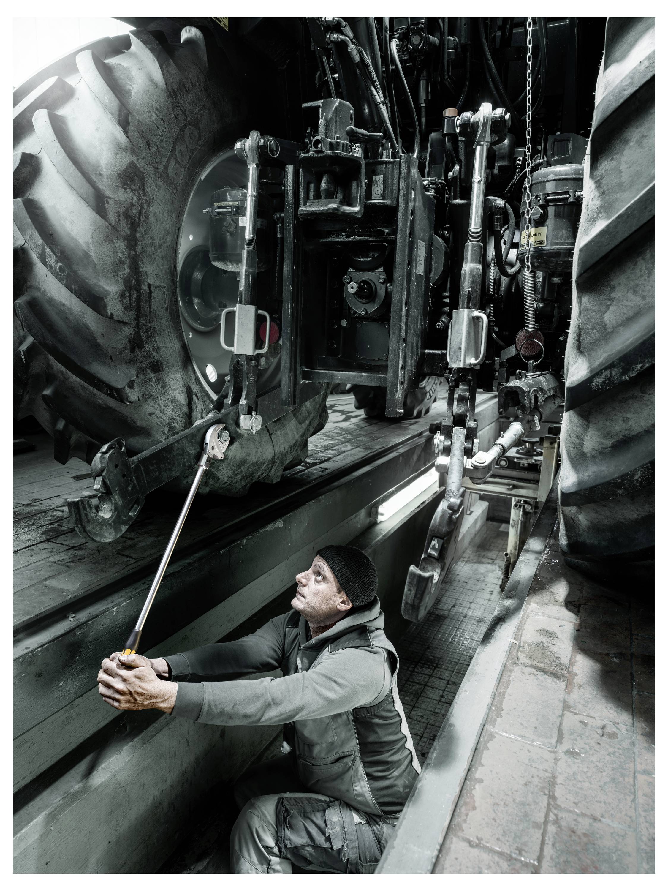 Technician crouches in a machinery trench, using a long tool to work beneath a large agricultural tractor.