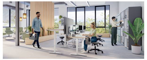 Modern open-plan office with employees working at desks, one man walking while carrying a bag, and another person organizing a cabinet near plants and large windows.