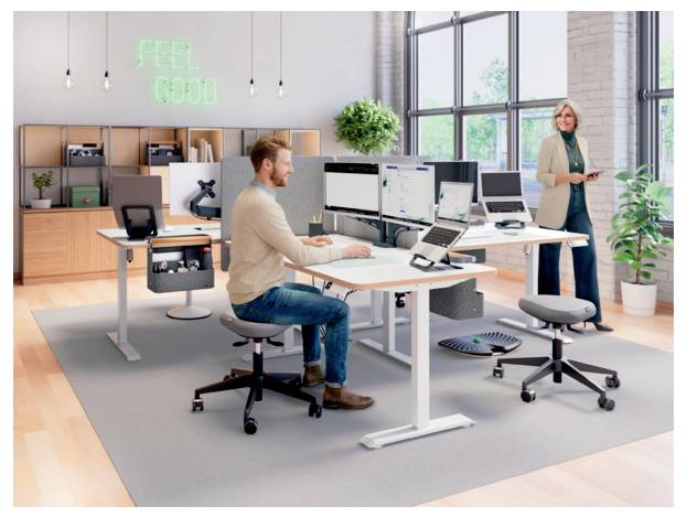 Modern open office with a man working at a standing desk and a woman nearby; neon sign reads 'FEEL GOOD'.