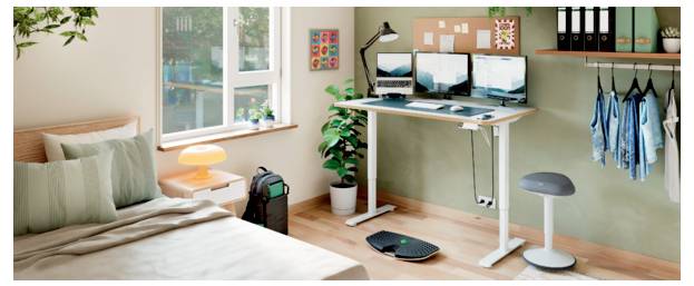 A small bedroom set up as a home office, with a bed beside a standing desk, dual monitors, chair, lamp, plant, and window.<br>