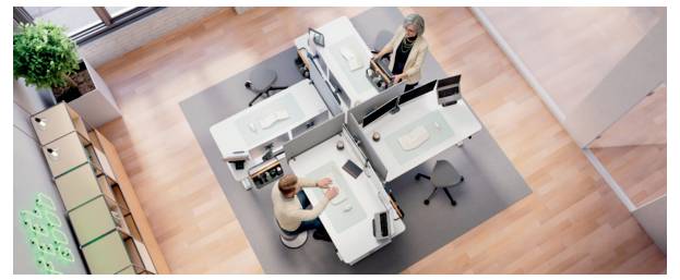 Overhead view of two people working at modular office desks in an open-plan workspace with partitions, stools, and a potted plant.