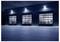Industrial warehouse exterior at night with three illuminated glass garage doors and a paved forecourt.