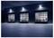 Nighttime exterior of an industrial warehouse with three lit garage doors and visible shelving inside behind a paved forecourt.