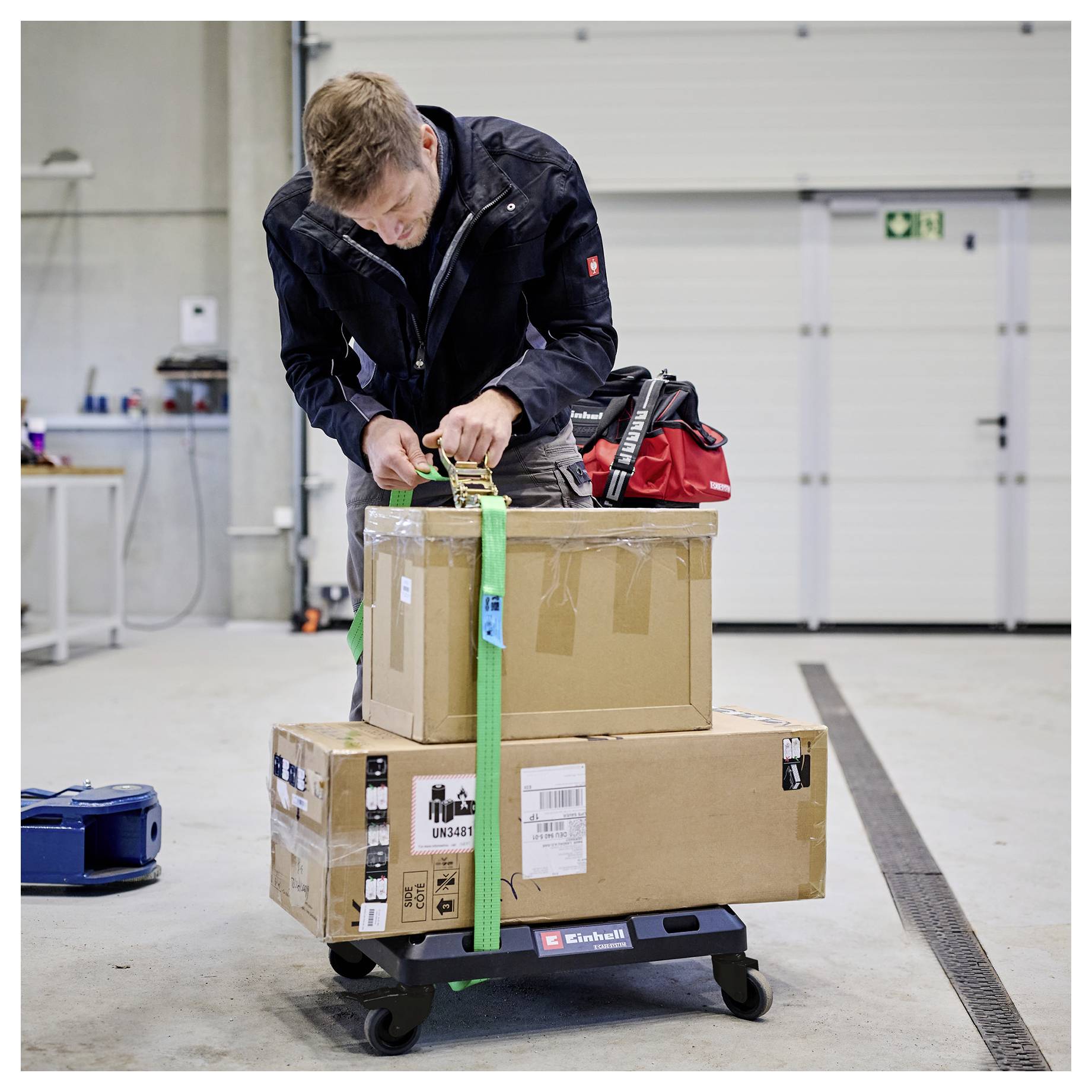 Worker straps a cardboard box to a wheeled dolly in a warehouse, preparing it for transport.