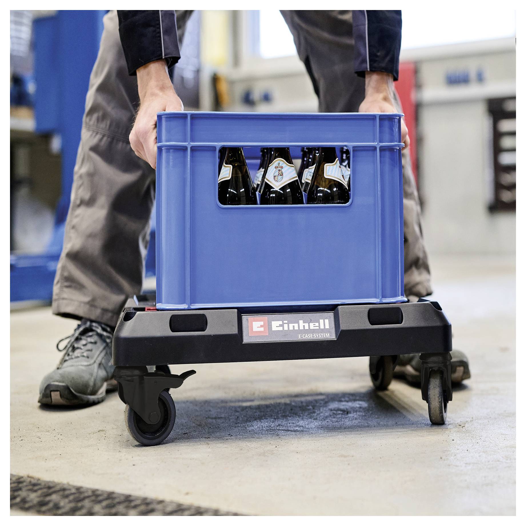 Worker pushes a blue crate of bottles on a wheeled Einhell cart in an industrial setting.
