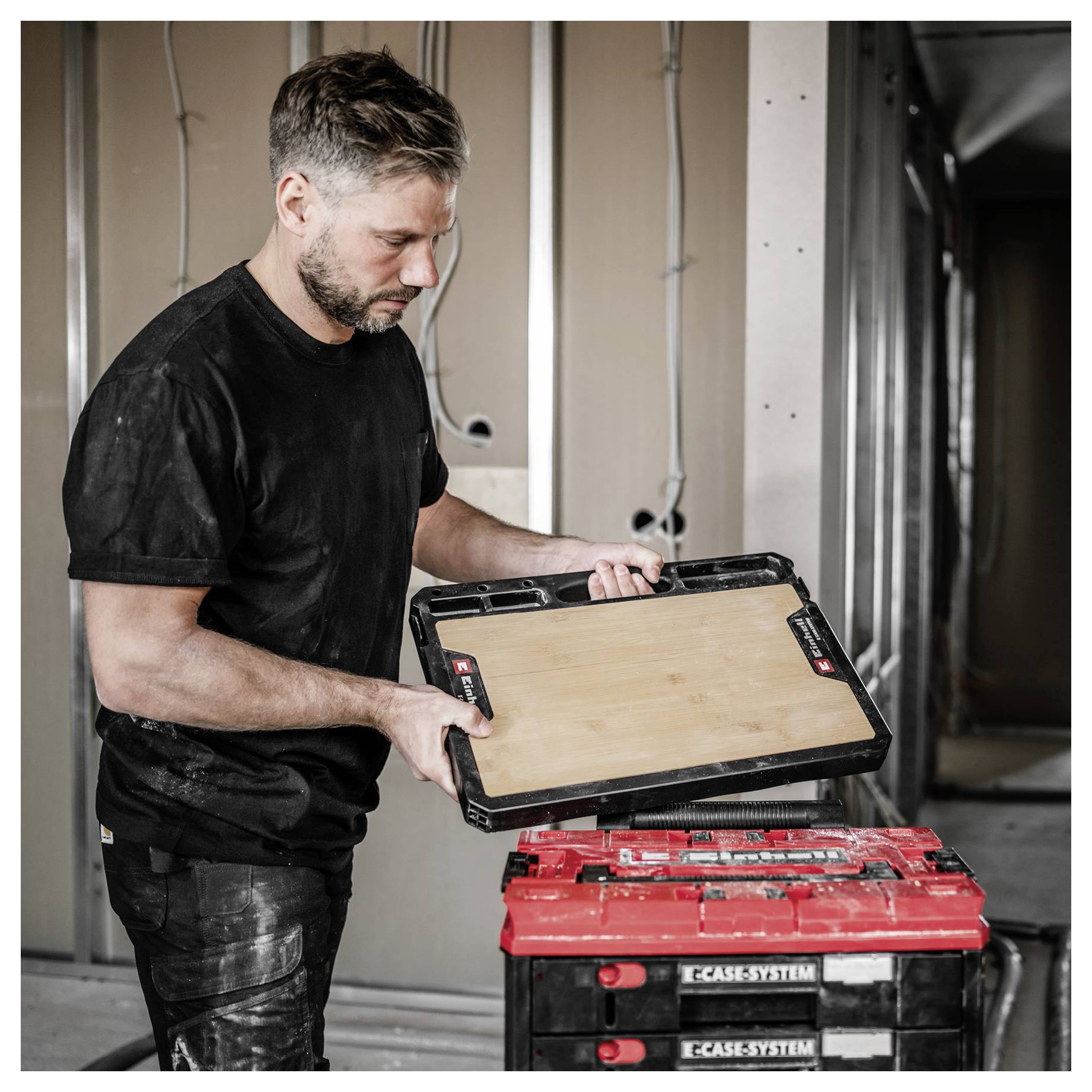 Construction worker fitting a wooden panel into a red-and-black CASESYSTEM toolbox or tool case stack indoors.