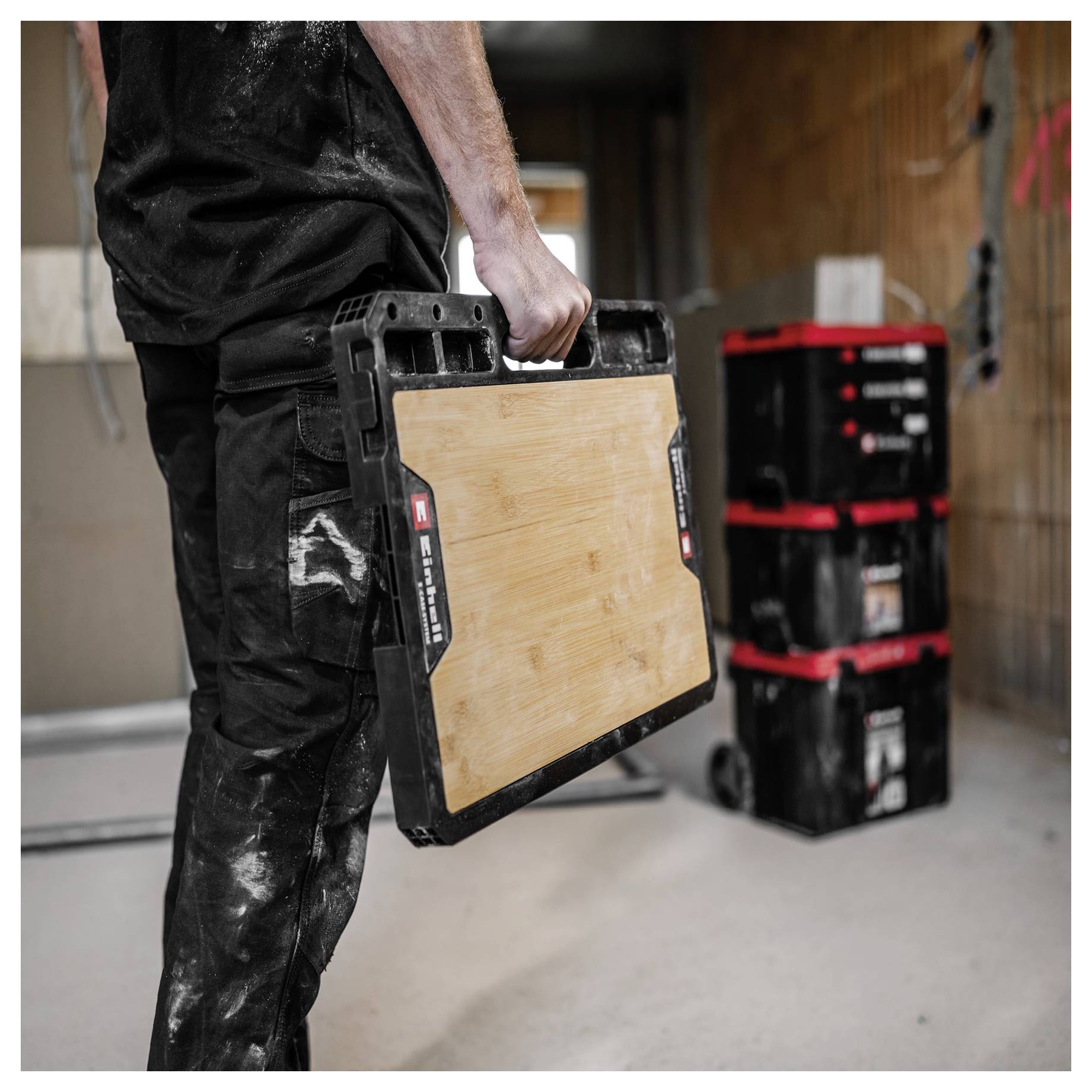 Construction worker carrying a folding work platform in a workshop, with stacked tool cases in the background.