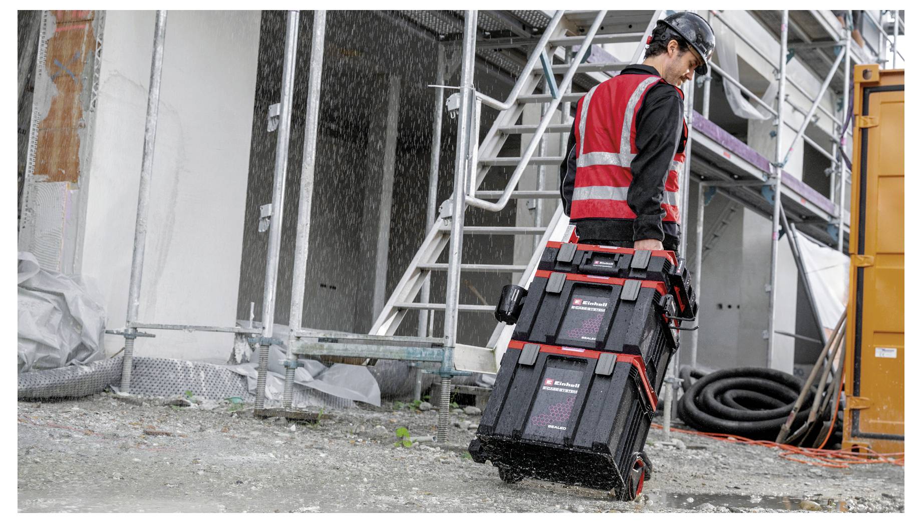 Construction worker in a red safety vest and hard hat carries stacked tool cases across a muddy worksite beside scaffolding and an orange container.