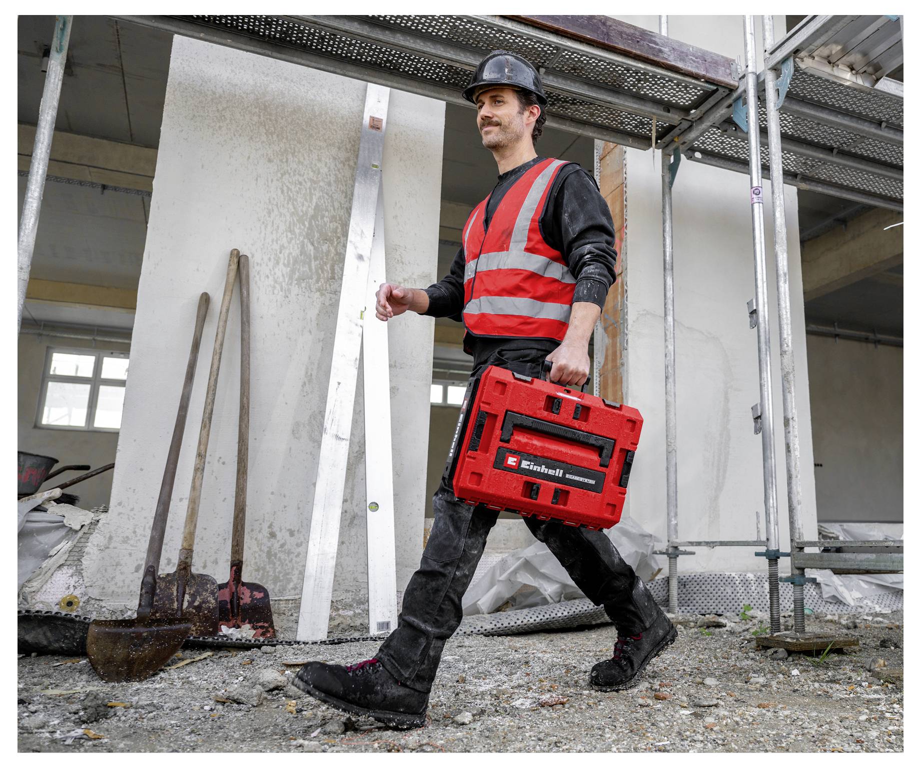 Construction worker in hard hat and safety vest walking with a red tool case on a building site.