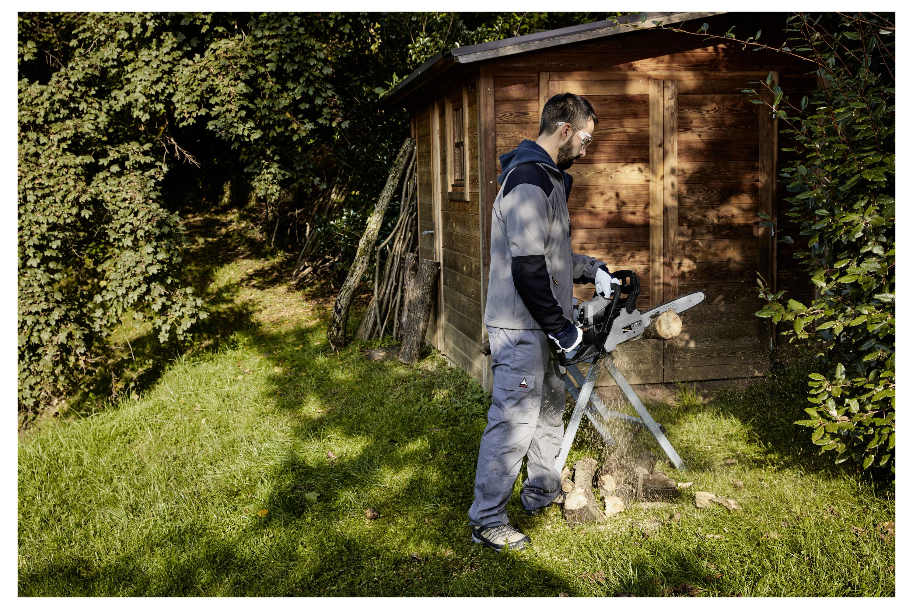Man operating a chainsaw near a wooden shed, cutting a log in a grassy yard with trees and sunlight.