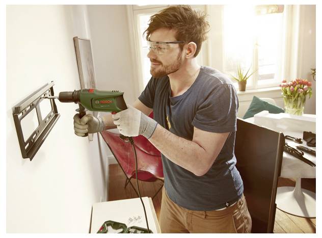 Man wearing safety glasses and gloves drills into a wall-mounted bracket with a power drill in a bright home interior.