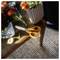 Orange-handled scissors on the edge of a wooden table beside a vase of flowers, with sunlight and a striped rug in the background.