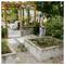 Woman rakes a raised garden bed beside a greenhouse, tending the soil in a backyard garden.