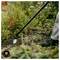 Gardener using a long-handled cultivator to weed and loosen soil around shrubs in a garden bed.
