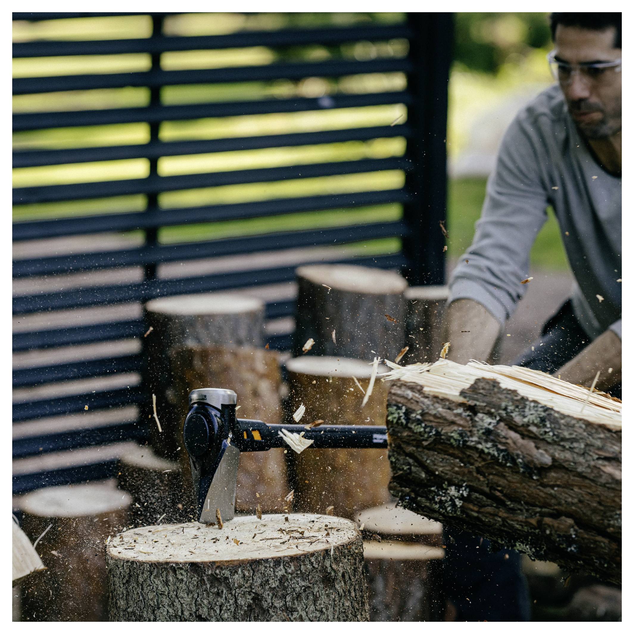 Man splitting a log with an axe outdoors, wood chips flying near stacked logs and a fence backdrop.