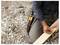 A person kneels outdoors on gravel, marking a wooden board with a red pencil while wearing a tool belt with hand tools.