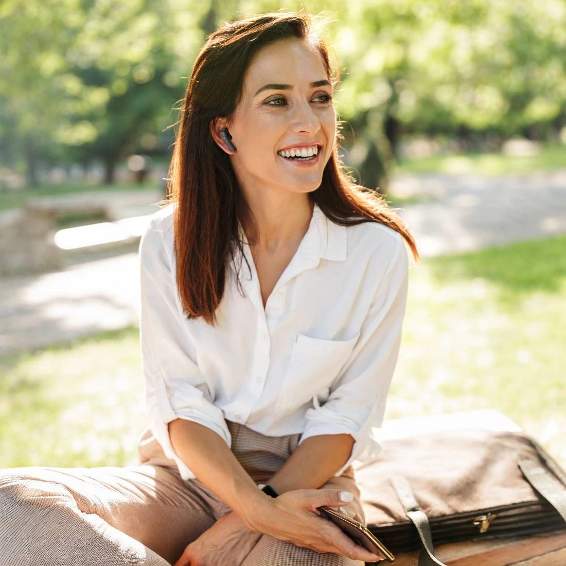 Smiling woman in a white shirt sitting outdoors on a bench in a sunny park, holding a phone and looking off to the side.