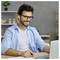 Smiling man wearing glasses and earbuds works on a laptop at a desk in a home office.