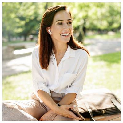 Smiling woman in a white shirt with earbuds sits outdoors on a park bench, holding a phone and looking off to the side.