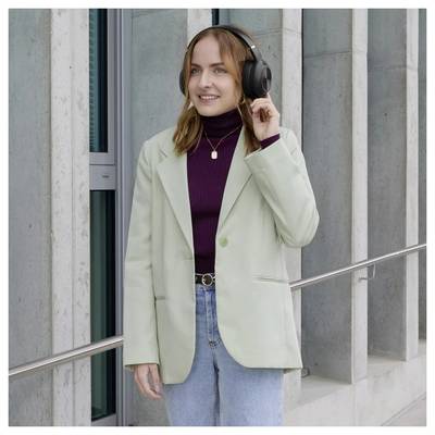 Woman wearing headphones and a light green blazer stands outdoors by a building, smiling and adjusting one earcup.