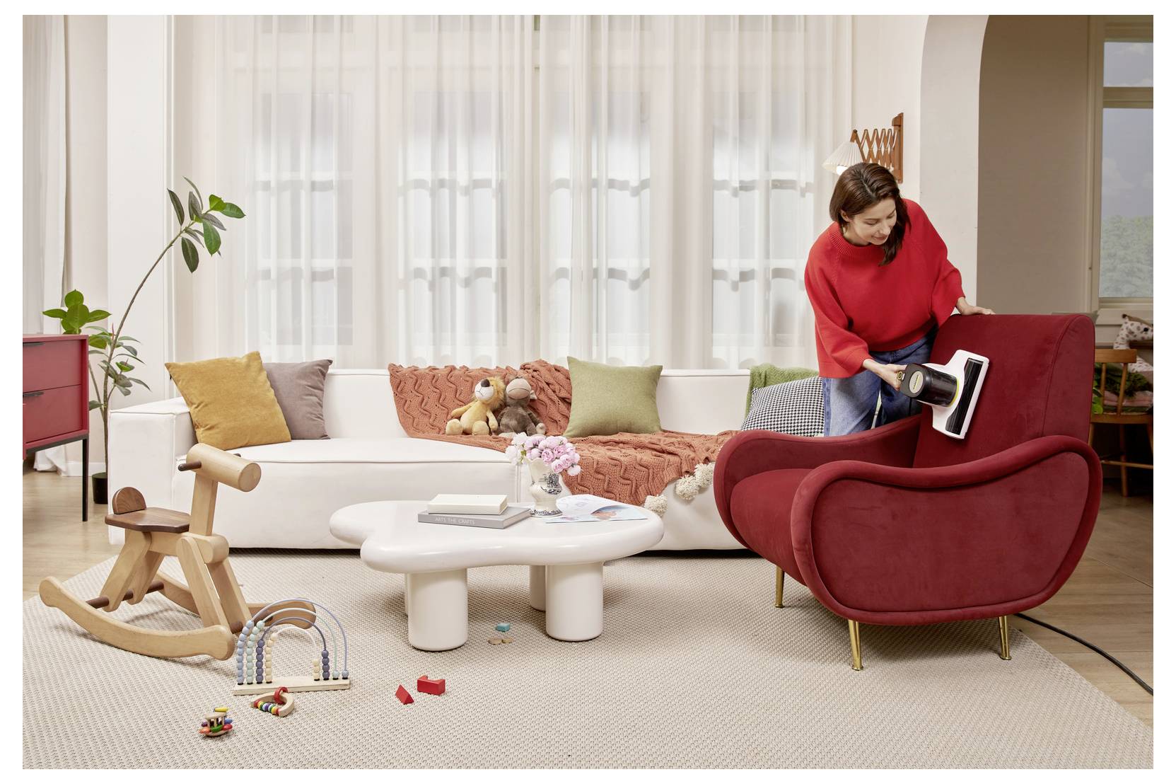 Woman vacuuming a red armchair in a bright living room with sofa, coffee table, toys on the floor, and a wooden rocker nearby.