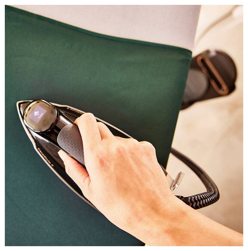 Hand holding a cordless iron over a dark green fabric, smoothing material on an ironing board.