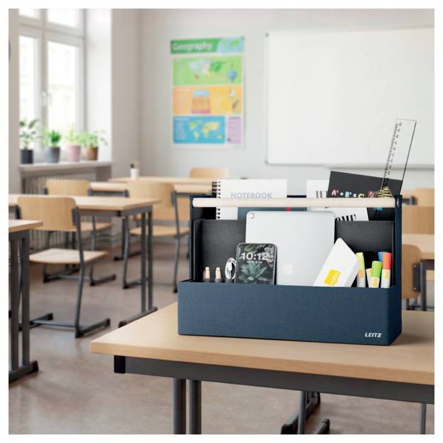 School classroom with empty desks and a desk organizer holding notebooks, pens, a tablet, and stationery in the foreground.