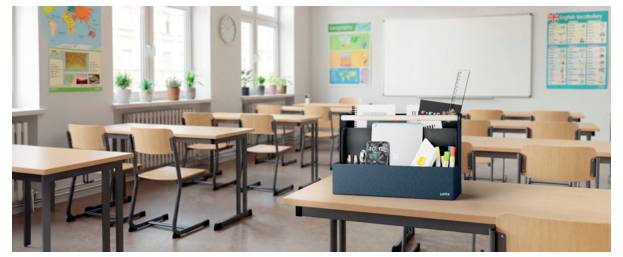 Empty classroom with rows of desks, a teacher’s desk, whiteboard, wall posters, and potted plants by the windows.