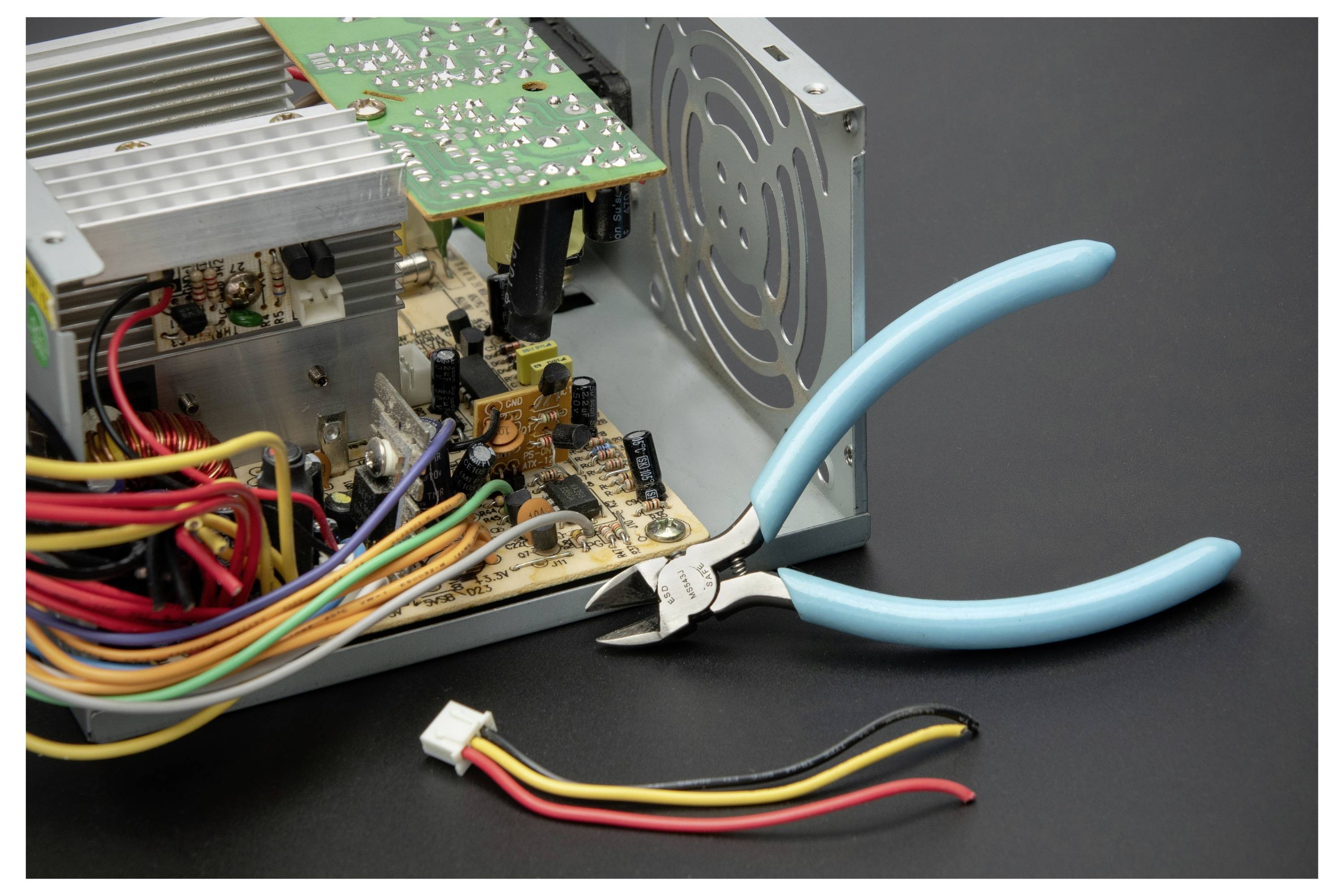 A computer power supply is open for repair, with exposed circuit boards, wires, a cooling fan, and blue-handled pliers beside it.