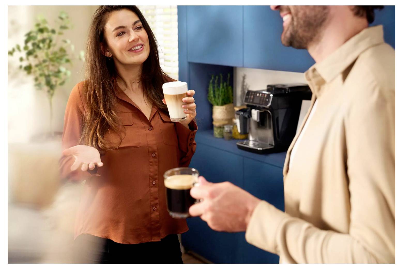 Two coworkers chatting in a kitchen, each holding a coffee cup, with a coffee maker and blue cabinets in the background.