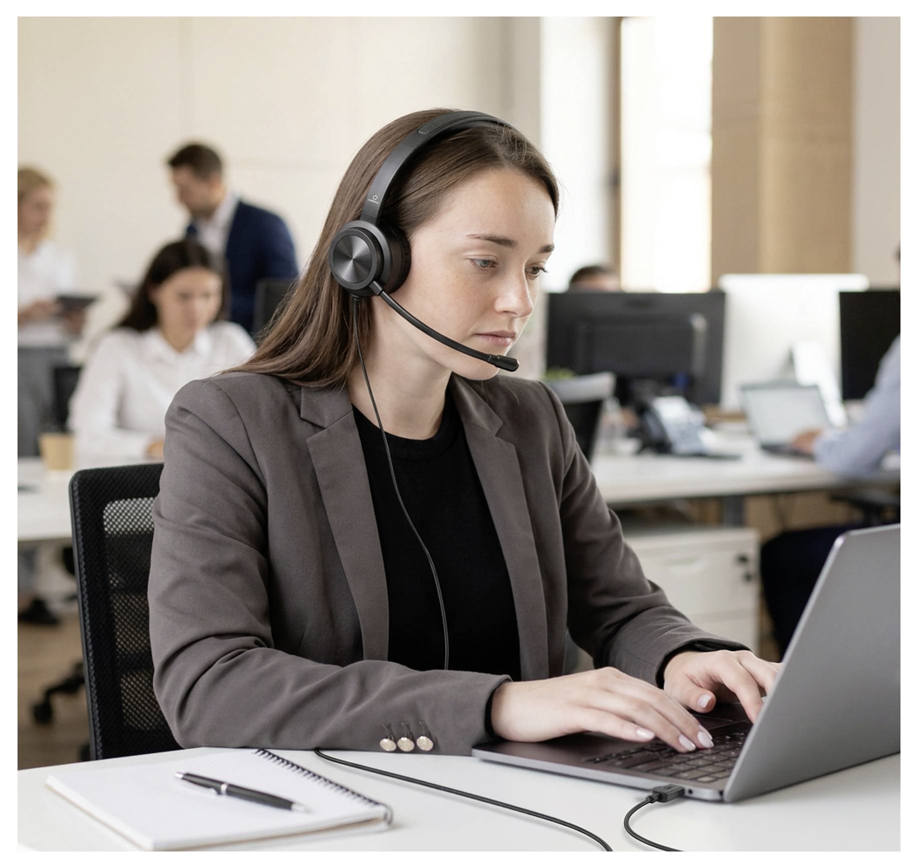 Woman wearing a headset works on a laptop in an office call center while colleagues sit blurred in the background.