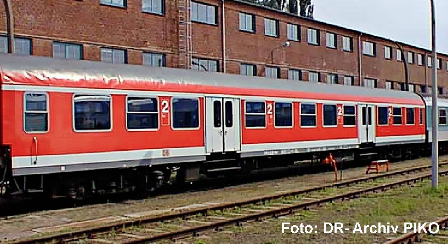 Red and white passenger train cars on railway tracks beside a brick building, with 'Foto: DR-Archiv PIKO' text in the corner.