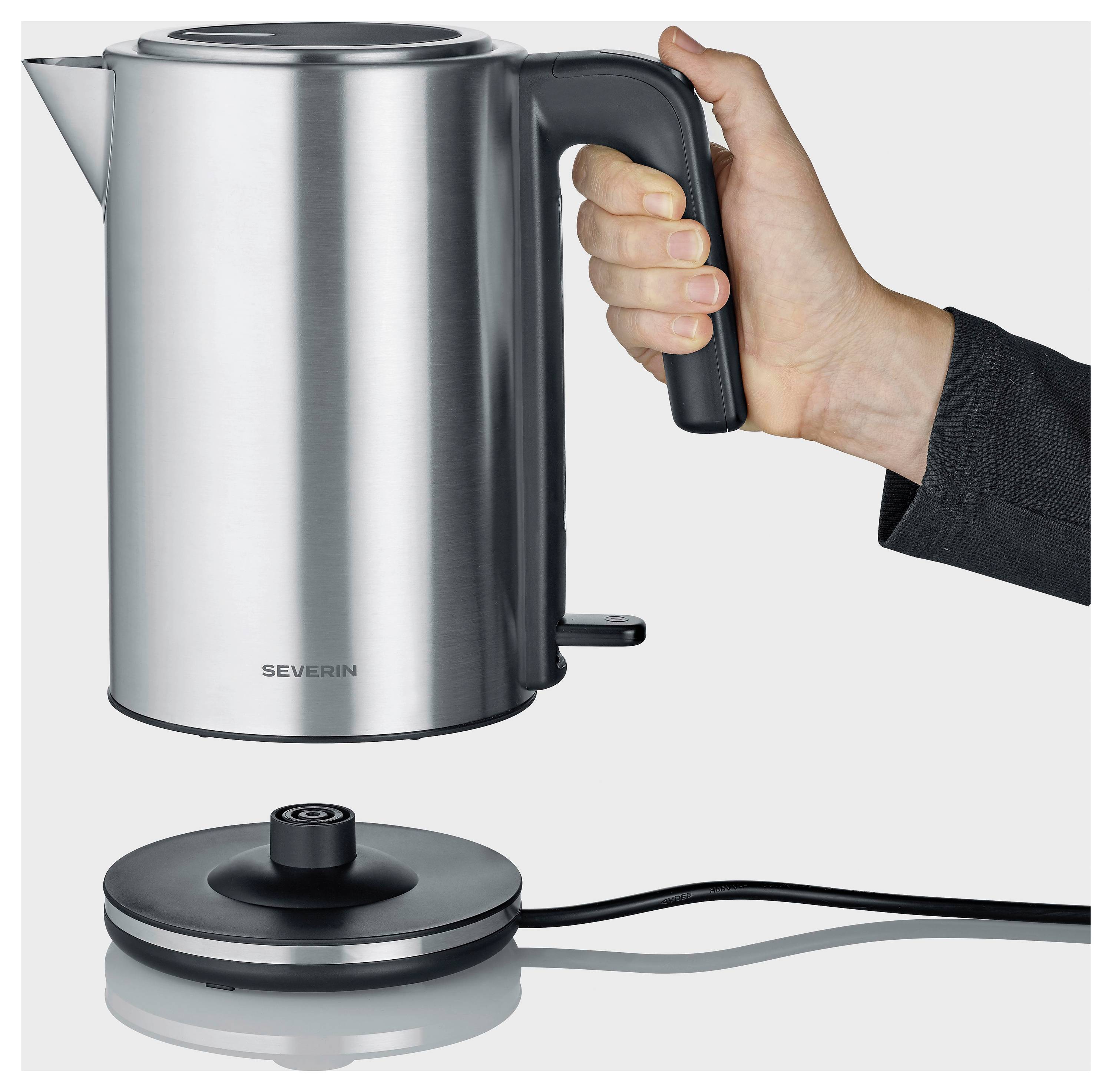 Hand holding a stainless steel electric kettle above its round power base with cord on a plain background.