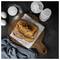 Freshly baked loaf of bread on parchment on a wooden board, surrounded by flour, eggs, sugar, and a bottle of milk on a dark countertop.