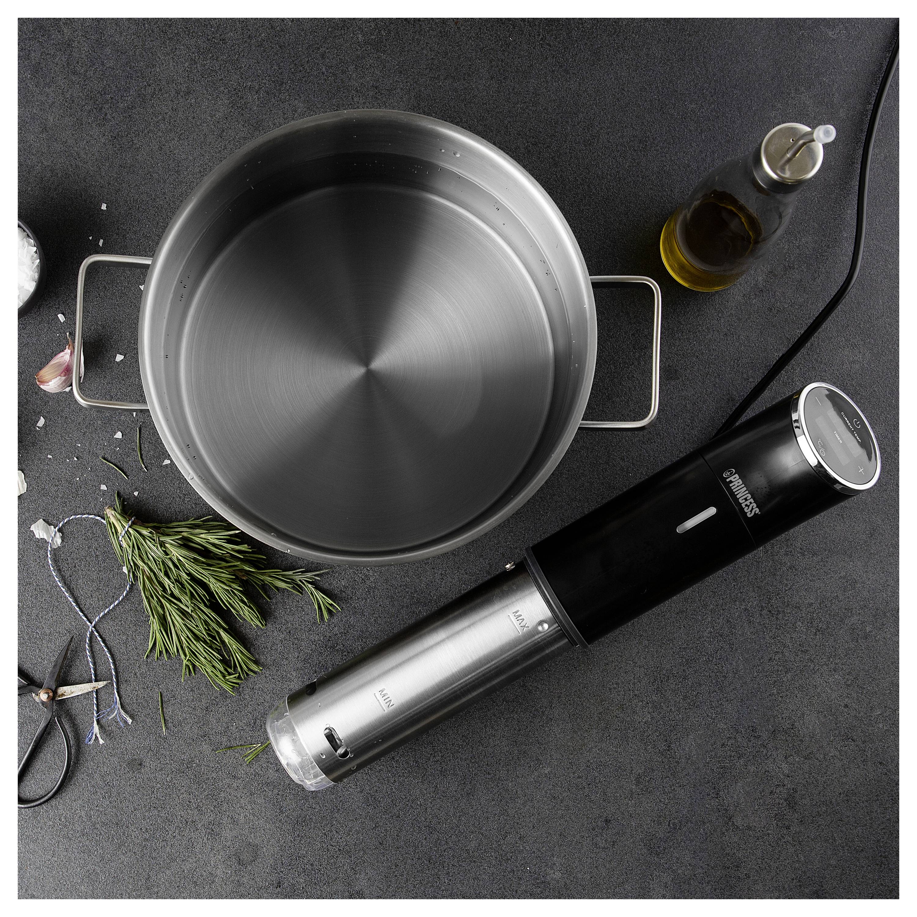 Top-down view of an empty stainless-steel pot with handles, a black-and-silver immersion blender, and a small bottle of oil on a dark countertop.