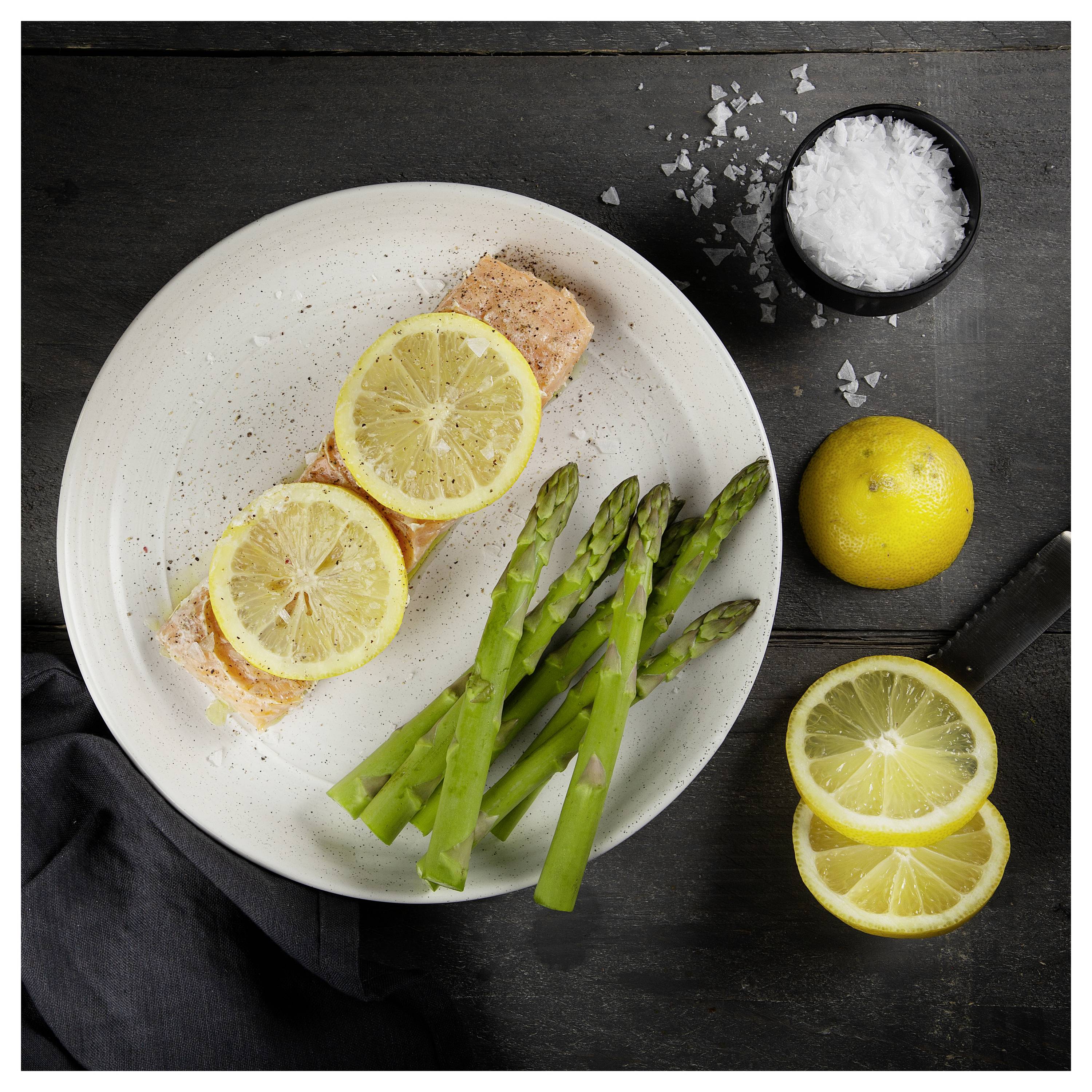 Plated salmon topped with lemon slices, served with asparagus on a dark tabletop beside whole lemons and sea salt.